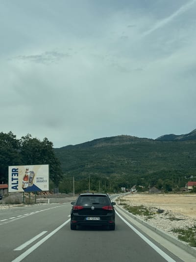 The image captures a view from inside a vehicle driving on a two-lane road in Jasenovo Polje, Montenegro. A black Volkswagen Golf, license plate NK DG676, is in the foreground, traveling in the same direction. To the left of the road, a large billboard displays an advertisement for "ALTER" beer, featuring two bottles and cans. The billboard is partially obscured by trees. The road curves gently to the right, leading towards a background of densely forested hills and mountains under a cloudy sky. On the right side of the road, there is an undeveloped area with dry grass and gravel, leading to scattered buildings. The scene suggests a rural or semi-rural setting with moderate vegetation and a vast, overcast sky.