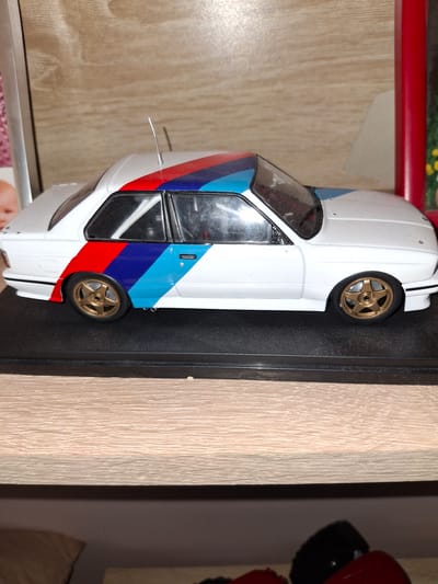 A model car, white with blue, red, and darker blue racing stripes, is displayed on a black base. The car has gold wheels and tinted windows. A thin, silver antenna extends from the rear of the car. The car is positioned on a shelf or display surface with a light-colored wood-grain texture. In the background, a red bar is visible above the shelf, and to the right, a blurred object with a red top and black bottom, possibly another collectible, is partially seen. The lighting suggests an indoor setting. The image appears to be taken from a slightly elevated angle looking down at the model car.