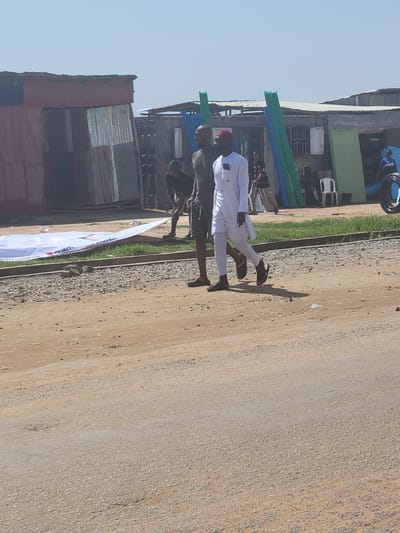 In Kwamba, Nigeria, two men walk on a dirt road adjacent to a row of makeshift structures constructed from corrugated metal. The man on the left wears a dark t-shirt and shorts, while the man on the right is dressed in a white traditional garment and a red cap. They are walking towards the right side of the frame. In the background, a large white banner with text is partially visible, lying on the ground. The structures in the background appear to be shops or informal dwellings, with various materials like metal sheets and tarps used for their construction. The sky is clear and bright, indicating a sunny day.