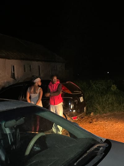 In Wumat, Nigeria, at night, two individuals, a woman and a man, stand near parked vehicles. The woman, facing away from the camera and to the left, wears a striped dress and a headscarf. The man, facing the camera and to the right, is dressed in a red vest over a dark shirt and white pants. They are positioned between a building with multiple windows and a dark-colored car. The foreground is occupied by the hood and windshield of another vehicle. The environment appears to be an outdoor, possibly rural setting, with vegetation visible in the background and a dirt ground surface. The scene is illuminated by artificial light sources, casting shadows and creating a dramatic atmosphere.