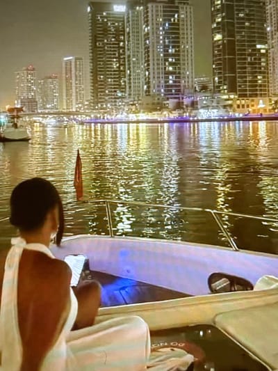 The image captures a nighttime view from the deck of a yacht, looking towards a cityscape across a body of water. Tall, illuminated skyscrapers dominate the skyline, their lights reflecting on the rippling surface of the water. A portion of a person, seen from behind, is seated on the yacht, facing away from the camera and towards the city. They are wearing a white garment that drapes off their shoulders, revealing their back. The person appears to be looking at a phone or tablet. A red flag is visible on a pole near the center of the frame. The yacht's interior, with plush seating and what appears to be underwater lighting, is also partially visible. The scene suggests a leisurely evening cruise or a moment of quiet contemplation against a vibrant urban backdrop. The image does not depict the city of Pori, Finland; instead, the architectural style of the skyscrapers suggests a location such as Dubai or a similar modern metropolis with a waterfront.