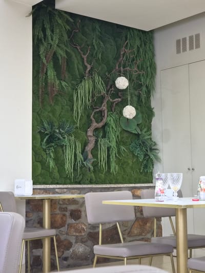 The image depicts an interior space, possibly a restaurant or dining area, showcasing a unique moss wall decoration. The wall is adorned with an arrangement of moss, ferns, and artificial branches, creating a miniature forest scene. Two white, spherical objects resembling flowers are suspended within the arrangement.

Adjacent to the moss wall is a dining area featuring a table with a light-colored surface and gold-colored legs. Surrounding the table are several chairs with light upholstery and gold-colored frames. The floor beneath the chairs is decorated with a stone pattern.

In the foreground, a small white object with printed text, possibly a branded item, is visible. The overall environment appears modern and well-maintained, blending natural elements with contemporary design.

The location of the image is Barcelos, Portugal.