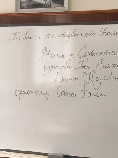 The image shows a whiteboard with text written in what appears to be Bulgarian. The text lists "Poems and Poems by Botev," followed by "Miguel de Cervantes," then the phrase "novel. Don Quixote," and finally "Alonso Quijano" and "armor-bearer Sancho Panza." The whiteboard is located within a room with a wooden panel and a framed picture on the wall. The bottom of the whiteboard has a grey eraser and what may be a cord. The image does not contain specific details that would allow the location to be confirmed as the city of Kavarna, Bulgaria.