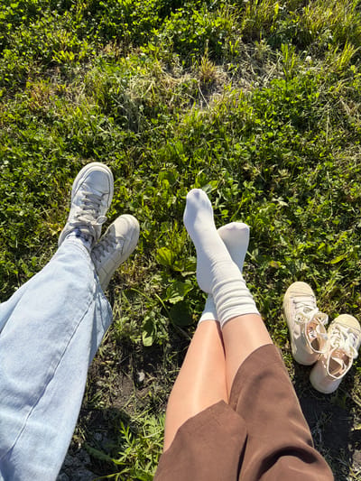 This is a top-down shot of two pairs of legs sitting on the grass in what appears to be a park or field in Yelizovo, Russia. One person is wearing light blue jeans, white sneakers with gray laces, and patterned socks. The other person is wearing a brown skirt and white ribbed socks, with a pair of beige canvas sneakers lying next to their feet. The ground is covered in green grass and some dry patches, suggesting an outdoor, possibly sunny, environment.