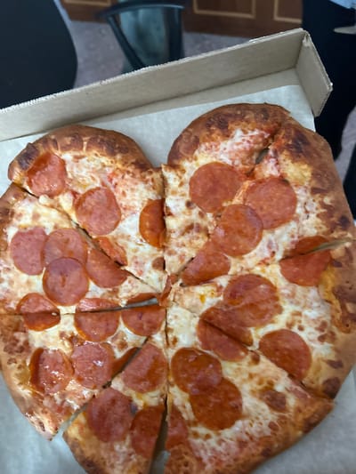 The image displays a heart-shaped pepperoni pizza contained within an open, light-brown cardboard pizza box. The pizza is generously topped with melted white cheese and numerous round slices of red pepperoni, exhibiting a slightly glossy appearance. The crust around the perimeter is visibly browned. The pizza is cut into multiple irregular triangular slices. The interior of the box features a white paper liner beneath the pizza. The background is out of focus, showing indistinct elements: a dark, possibly metallic or plastic object on the left and a blurred dark surface, potentially wood or furniture, on the right. No individuals, specific actions, or interactions are discernible. The environment appears to be an indoor setting with ambient lighting.