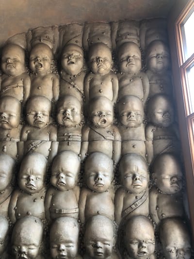 The image depicts a sculptural wall relief, featuring numerous casts of infant figures. They are arranged in rows, appearing densely packed and slightly overlapping. The infants are depicted lying supine, with closed or partially open mouths, suggesting a state of repose or possible distress. Some are adorned with what seems to be a band or strap across their bodies. The texture of the sculpture is rough, with small nodules dispersed across the surface.

The location of the sculpture is Epagny, Switzerland. The work is likely situated indoors, evident by the natural light from an unseen window or skylight at the top of the image.