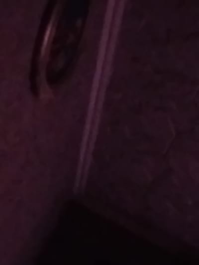 A close-up, blurry, and dimly lit image shows a portion of a wall with a textured pattern, possibly hexagonal, in a dark purple hue. A vertical, lighter purple line runs down the right side of the image, suggesting a corner or a seam. In the upper left corner, a circular object with a metallic rim is visible, partially obscured and out of focus. It is impossible to determine the exact nature of this object or its surroundings with the given visual information. The image does not contain any discernible features related to people, actions, or interactions.
