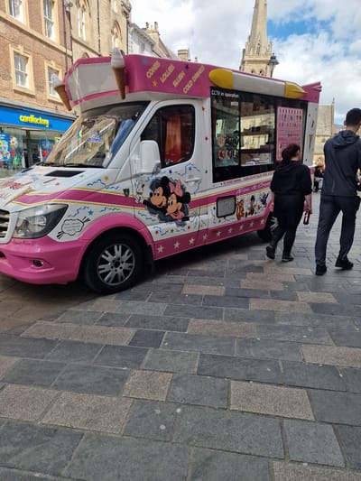 Here is a concise description of the image:
The image shows a pink and white ice cream van parked on a brick-paved street in Durham, United Kingdom. Two people stand near the van. The van's design includes images of Disney characters. Markings on the van state "Princess Maxine VII", "Static Site And Event Operator", "Fleet No. 10", "CONES", "LOLLIES", "SLUSH", "COLD DRINKS", and "CCTV in operation". A cardfactory store is visible behind the van. A gothic church spire rises in the background.