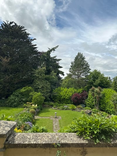 The image depicts a lush, green garden with a stone wall in the foreground. A neatly mown lawn is visible, bordered by a variety of trees, shrubs, and flowering plants. A stone pathway cuts across the lawn, leading towards a small stone feature. The garden is surrounded by dense foliage and mature trees, suggesting a well-established landscape. The sky above is partly cloudy, with a mix of blue sky and white clouds. The scene is peaceful and idyllic, evoking a sense of tranquility. The location is the botanical gardens in Clondalkin, Ireland.