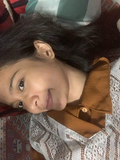 The image shows a close-up of a young woman's face as she lies down, possibly on a bed, in a room. Her head is tilted, and she is looking towards the viewer with a slight smile. Her dark hair is spread out around her. She is wearing a brown collared shirt with a white lace overlay. The background appears to be a patterned blanket or rug, with hints of red, brown, and cream colors. A green and white striped pillow is visible behind her head. There is no indication of the specific location within Sungai Meriam, Indonesia.