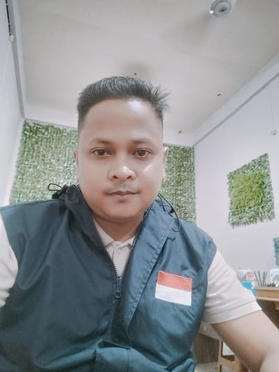 The image shows a selfie of a man in an indoor setting. He is looking directly at the camera, with a neutral expression. He is wearing a dark blue vest over a light-colored collared shirt. On the left chest of the vest, there is a patch with the Indonesian flag, which is red and white. In the background, there are walls with artificial green foliage panels. To the right, a portion of a wooden desk and a plastic water jug are visible. The lighting suggests it is daytime.