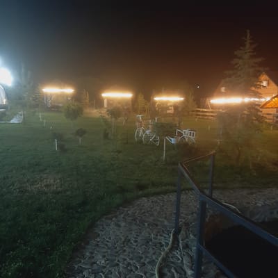 This nighttime photograph captures a courtyard or garden area illuminated by warm outdoor lighting. The scene features several elements typical of an outdoor gathering space or recreational area. In the foreground, a paved walkway made of dark, irregular stones curves into the frame, bordered by a dark metal railing. Beyond the walkway, a lush green lawn stretches out, dotted with small trees or bushes. Two decorative metal bicycles, one appearing more elaborate than the other, are placed on the lawn, suggesting a whimsical or rustic aesthetic. Further back, several gazebos or covered seating areas are visible, each topped with bright, linear lights that cast a warm glow. A tall evergreen tree stands to the right of the frame, with a portion of a building, possibly a house or a lodge, visible behind it, also illuminated. The sky is dark, indicating nighttime, and the overall atmosphere is tranquil and inviting. The location is identified as being in Sosnovka, Kyrgyzstan.