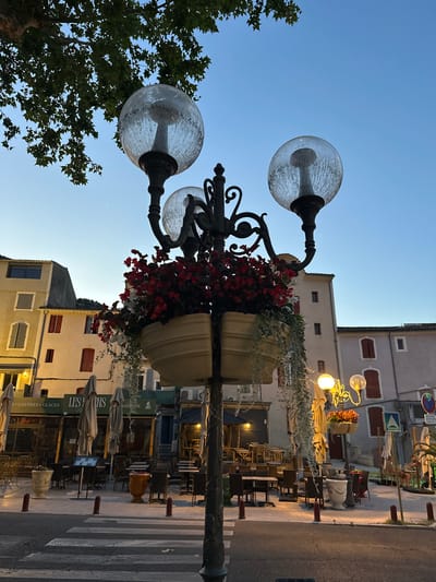 The image depicts a street scene in Anduze, France, at dusk. A lamppost with three glass globes and a planter filled with red and white flowers stands in the foreground. Behind the lamppost, buildings with closed shutters line a street where outdoor seating for restaurants is set up. A crosswalk is visible in the lower part of the image. The sky is a clear twilight blue.