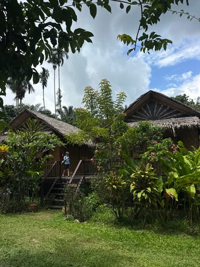 A boy in a blue shirt and dark shorts stands on a wooden staircase leading up to a thatched-roof bungalow in Tambon Khlong Thom Nuea, Thailand. The bungalow has walls made of woven bamboo and a wooden deck. Lush green foliage, including palm trees, surrounds the structure. The sky above is partly cloudy, with patches of blue visible. The scene appears to be a peaceful, tropical setting.