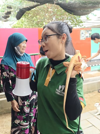 In an outdoor setting, possibly a zoo or farm, a young woman wearing glasses and a green polo shirt is holding an orange iguana. The woman's hair is pulled back, and she is wearing a black undershirt. She appears to be speaking, with her mouth open and a surprised or excited expression. She is also holding a red and white megaphone in her left hand. Behind her, an older woman in a blue hijab and a black floral dress is visible, looking to the side. In the background, another person, a young male, is partially visible. The scene is outdoors with foliage and a netted roof overhead. The ground appears to be a dirt or sandy surface with some paved areas. A white rabbit is also visible in the lower right portion of the image. The text "TIENTIEN FARM & ZOO" is visible on the woman's green shirt. The image suggests a daytime scene, likely warm given the outdoor setting. The overall mood seems to be educational or informational, perhaps a guided tour or presentation.