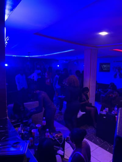 This is a nighttime, indoor photograph taken in what appears to be a dimly lit lounge or club in Wumat, Nigeria. The scene is dominated by vibrant blue and red lighting, casting a moody ambiance. Several individuals, predominantly young adults, are present and engaged in various activities.
In the foreground, a person in a dark top is bent over a table filled with drinks and other items. To the left, a counter area displays containers and utensils, suggesting food or beverage service.
The midground features a group of people gathered around couches and seating areas. Some are socializing, while others appear to be resting or observing the surroundings. A prominent striped column is visible in the center of the room.
In the background, more individuals are seen, some standing and others seated. Artwork or murals adorn the walls, with a "NO SMOKING" sign also visible. The ceiling is illuminated with strip lighting and a central fixture. The overall impression is that of a social gathering at an establishment that offers entertainment and refreshments.