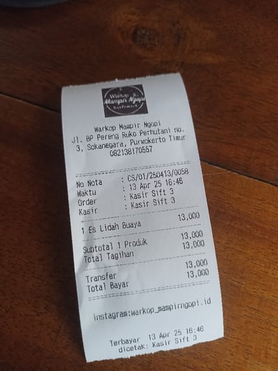 The image shows a receipt from a food stall called "Warkop Mampir Ngopi" located at Jl. BP Pereng Ruko Perhutani no. 3, Sokanegara, Purwokerto Timur, Indonesia. The phone number of the stall is 082138170557. The receipt was issued on April 13th, 2023 at 16:46, with order number CS/01/250413/0058, and was served by cashier shift 3. The receipt lists one item "1 Es Lidah Buaya" for 13,000 Indonesian Rupiah. The total bill was 13,000 Indonesian Rupiah, and the customer paid with a transfer. The receipt has an Instagram handle for the stall: "warkop_mampirngopi". The receipt was printed at 16:46 by cashier shift 3. The image is likely taken in a food stall or restaurant. The receipt is lying on a wooden surface.