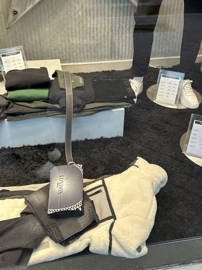 This media file captures a close-up view of a shop window display, likely in Koridallos, Greece, showcasing a selection of contemporary clothing and accessories. The scene is dominated by a black, shaggy rug that serves as a backdrop for the merchandise.
The primary subjects include several clothing items. On the right, a cream or off-white sherpa-style jacket with dark trim is prominently displayed. Beside it are folded dark-wash jeans. Towards the center and left, a stack of folded garments includes a black item, a vivid green item, and an olive-green patterned jacket or blazer. A long, thin black and white striped accessory, possibly a clutch bag or wallet, rests on the rug, featuring the text "LONDON NOVI" in a distinctive radial font.
Other objects in the display include two clear acrylic stands holding white paper signs that appear to list product details, likely sizes and prices. One sign is positioned next to a pair of white chunky-soled sneakers, which are placed on a small, metallic stand. Another set of white sneakers is visible on the lower left, accompanied by another price list. A rectangular white plinth or box is situated among the folded clothes.
The activity depicted is the merchandising of fashion items in a retail setting. The display is designed to attract customers, and the presence of reflections on the glass indicates someone observing the window. A notable detail is the clear reflection of a person's lower leg and foot, wearing black trousers and white sneakers, suggesting a passerby or potential customer. The reflected environment outside the shop features a patterned floor or wall.
Visible text includes "LONDON NOVI" on the striped accessory. The signs in the acrylic stands contain text that is somewhat obscured by reflection and blur, but appears to list product information, potentially sizes like "170 cm" and prices in euros, such as "27€" and "29€." The top of one sign seemingly reads "MAJI LEKI." The overall color palette of the display is largely monochromatic with black, white, and grey tones, accented by the olive green and brighter green clothing items.
