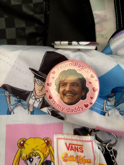 A close-up shot shows a pink circular pin with a picture of Pedro Pascal on it, surrounded by small red hearts. The text on the pin reads "Pedro Pascal is my daddy". The pin is resting on a white fabric with a pattern of anime characters. To the left of the pin, a character wearing a tuxedo and top hat is visible. Below the pin and anime characters, another fabric is visible with a yellow anime character on it, and a white label with "VANS Sailor Moon" printed on it. A metal keychain ring is attached to the right side of the label. The background appears to be a black and white checkered pattern. The image does not provide any information about the city of Červená Píska, Czechia, or any specific location within it.
