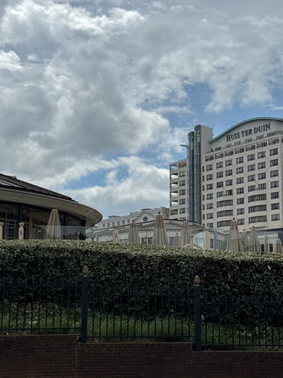 The image, when correctly oriented, presents a daytime view of the Hotel Huis ter Duin in Noordwijk, Netherlands, under a partly cloudy sky. The main subject is the tall, light-colored hotel building, prominently displaying "HUIS TER DUIN" in large letters along its distinctive curved roofline on the left side of the frame. The building features many floors with a regular pattern of rectangular windows and some balcony areas.
In the mid-ground, a lush, dark green hedge runs horizontally, bordered by a black metal fence. Situated between this hedge and the hotel facade are several light-colored patio umbrellas, indicating an outdoor dining or seating area. Another, less defined building is partially visible behind the umbrellas on the right side. The foreground on the right consists of a dark brick wall or path. The sky above is a mix of white and grey cumulus clouds interspersed with patches of clear blue, suggesting fair weather. There are no people or discernible activities taking place in this static scene.