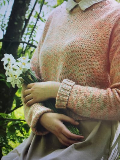 The image shows a close-up of a person wearing a knitted sweater and holding a bouquet of white flowers. The sweater is a speckled pink and orange color. The person's hands are visible, and they are gently holding the stems of the flowers. The background is out of focus and appears to be a natural outdoor setting with green foliage and sunlight filtering through. There is no identifiable location in the city of Cherepovets, Russia.