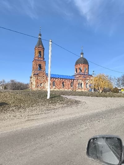 The image displays a two-part brick church, identified as located in Oktyabr'skoye, Russia, under a clear blue sky with sparse clouds. The structure features a tall, weathered reddish-brown brick bell tower on the left with a dark, conical spire topped by a finial, and arched openings. To its right, the main church body, also of reddish-brown brick, is topped by a large dark dome with a cross finial. The roof sections connecting the tower and the main body are a distinct blue. The church facade exhibits multiple arched windows.

In the foreground, a paved road surface occupies the lower right, transitioning to a dirt or gravel path on the lower left. A patch of dry, sparse grass separates the road from the elevated ground supporting the church. A white utility pole rises vertically in the mid-foreground, with two dark power lines extending horizontally across the upper portion of the frame. Bare-branched trees are visible to the left and behind the church, while a tree with vibrant yellow autumn foliage stands to the right. The lower right corner of the image includes the frame of a dusty, dark vehicle side-view mirror, reflecting a blurred interior element. No people are present.