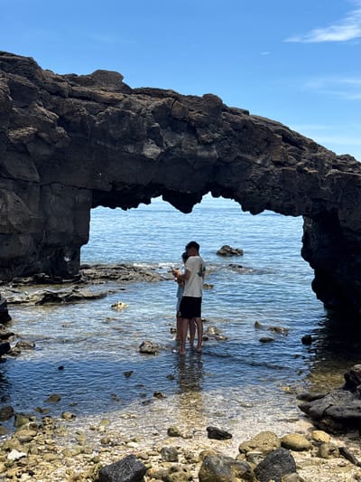 In Lý Sơn Special Administrative Region, Vietnam, a couple stands in shallow, clear ocean water under a natural rock archway. The archway is made of dark, rough-textured volcanic rock, with the sea visible through its opening. The water is calm and reflects the bright blue sky. The foreground is a rocky shoreline with a mix of dark volcanic stones and lighter, weathered rocks. The couple, facing away from the camera, appears to be embracing or interacting closely. The man is wearing a white t-shirt with a graphic print and black shorts, while the woman is in a light-colored dress or top. Their feet are submerged in the water. The overall scene depicts a serene moment at a coastal geological formation.