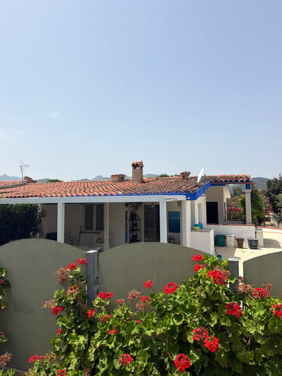 The image shows a sunny exterior view of a white house with a red tile roof and blue trim, situated in Santu Diadòru/San Teodoro, Italy. In the foreground, a low, curved, light green wall partially obscures a dense cluster of vibrant red geraniums in full bloom. The house features an open-air covered patio with white pillars and a white roof extending from the main structure. A window and a doorway with a blue frame are visible on the ground floor of the house. Behind the patio, a second, slightly taller section of the house is visible, also with a red tile roof and blue trim. A satellite dish is mounted on this section of the roof. Several trash bins (green, white, brown, and grey) are placed on a light-colored paved area near the house. In the background, distant hills and mountains are visible under a clear, pale blue sky. An antenna is mounted on the roof of the main house section. The overall scene depicts a residential area on a bright, clear day.