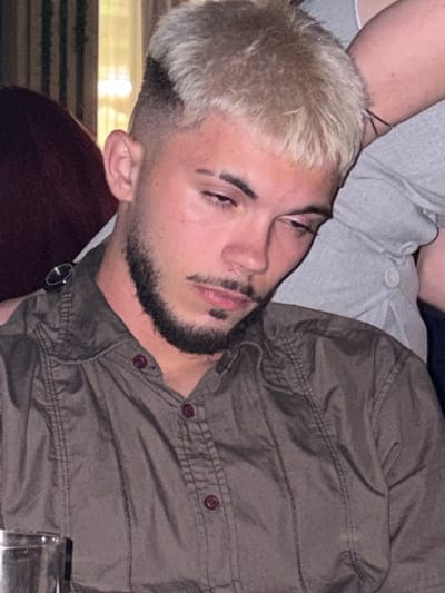 A young adult male with bleached blonde, textured hair and dark, faded sides is the central figure, leaning his head slightly to his right (viewer's left). He has a dark, well-groomed beard and mustache, and prominent dark eyebrows. His eyes are cast downwards, appearing relaxed or introspective. He wears a long-sleeved, collared shirt in a muted olive-brown color, featuring vertical stitched seams on the chest and dark reddish-brown buttons.
To his immediate right, the arm and upper torso of another individual in a light grey, possibly ribbed, casual top are visible, with the arm resting gently over the central figure's head/shoulder. Two light-colored buttons are visible on the grey garment. To his left, a portion of a third person wearing dark maroon or red clothing is visible. A small, dark-faced wristwatch with a dark strap rests on the central figure's left shoulder, appearing to belong to the person in maroon. In the bottom left corner, the metallic rim of a drinking glass is partially visible.
The background suggests an indoor setting with ambient lighting, possibly a restaurant or social gathering space, with a hint of a window or light source behind vertical blinds in the upper left. The scene depicts a moment of casual interaction and relaxation in Slivnitsa, Bulgaria.