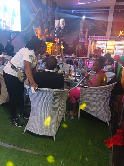 The photo was taken in an entertainment venue in Syokimau-Mulolongo ward, Kenya. The floor is covered with artificial grass. The furniture is primarily white wicker. A woman wearing a white t-shirt with a "MOK" logo and black pants interacts with a man wearing a black shirt. A woman in a pink outfit sits across from them, smoking a hookah. Hookahs and drinks sit on the table. A bar with illuminated shelves and bottles of alcohol is visible in the background. A mural decorates the wall. Other patrons are present.