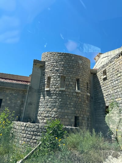 The image shows a stone tower in Donji Katun, Montenegro. The tower is cylindrical with rectangular window openings. Adjacent to the tower is a stone building. Vegetation, including bushes and trees, grows around the base of the tower and building. The sky is bright blue. The image appears to be taken through glass, as evidenced by reflections.