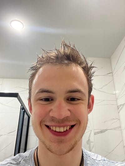 A close-up, frontal selfie captures a light-skinned young adult male smiling broadly directly at the camera. His short, brown hair is wet and styled with a spiky appearance, showing small droplets of moisture. He exhibits light stubble on his chin and jawline. He wears a light-colored, possibly striped, shirt and a dark necklace is visible around his neck. The background consists of a modern bathroom interior. White or light-grey large format tiles with subtle grey veining cover the walls. A portion of a black-framed shower enclosure is visible on the left. The ceiling is plain white, featuring a single, circular, recessed LED light fixture illuminating the scene. The specific city of Tliustenkhabl', Russia, cannot be determined from the visual information provided; however, the scene depicts an indoor bathroom setting.