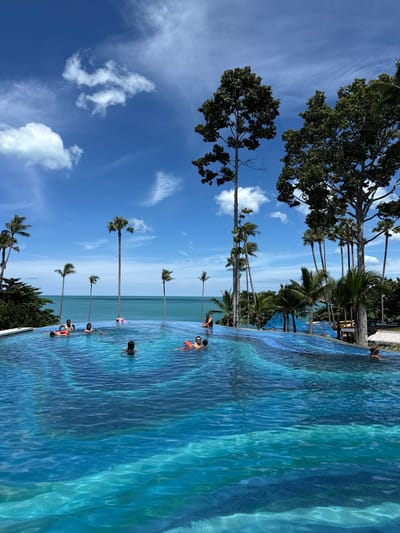 The image depicts an infinity pool in Choeng Mon, Thailand, with a clear blue sky and fluffy white clouds. The pool is filled with vibrant turquoise water, and its edge seamlessly merges with the ocean in the background. Several people are enjoying the pool, some swimming, others relaxing on inflatable tubes, and a few are conversing. Palm trees and lush green foliage surround the pool area, with tall, slender trees standing prominently against the sky. The scene conveys a sense of relaxation and tropical vacation.