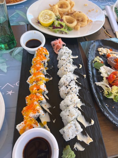 The image depicts a table setting at a restaurant in San Roque, Spain, featuring various dishes of food. In the foreground, a black rectangular platter holds two rows of sushi rolls. The sushi on the left is topped with bright orange roe and a yellowish sauce, while the sushi on the right is coated with sesame seeds and drizzled with a white sauce. To the left of the platter is a small white dish containing soy sauce, and to the right, a small mound of green wasabi. In the background, a white plate holds fried calamari rings and a side salad with a lemon wedge. Another black plate to the right contains a portion of tuna sashimi with a garnish of lettuce and cherry tomatoes. A green glass bottle, likely containing a beverage, is visible on the left side of the image. The table itself is made of wood, and a blue placemat with a tropical leaf pattern is visible beneath some of the dishes. The scene suggests a meal is in progress, with no people clearly visible.