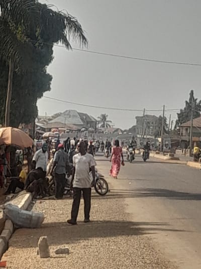 This image depicts a bustling street scene in Ube, Nigeria, during daytime with a hazy sky. The primary focus is a two-lane asphalt road. On the left side, an unpaved shoulder transitions into a pedestrian area containing concrete blocks, bagged materials, and a light brown umbrella providing shade over a gathering of individuals. An individual in a white short-sleeved shirt and dark trousers stands prominently in the foreground, facing the camera and holding a light-colored object. To their left, several men are congregated; one wears a white shirt and green cap, while another in a yellow t-shirt is seated.
Further along the asphalt road, a woman in a pink patterned dress walks away from the camera. Multiple motorcycles, some with riders and passengers, are present on the road, indicating active vehicular traffic. In the background, various single and multi-story buildings with corrugated metal roofs line the street. Dense tropical foliage, including palm trees, is visible on both sides of the road. Overhead power lines traverse the upper portion of the frame. Shadows are prominent, suggesting a bright, possibly mid-morning or afternoon sun. The overall environment suggests a dynamic urban or semi-urban setting.
