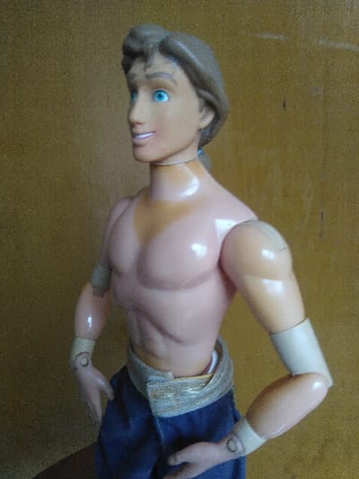 A mid-shot photograph, taken in Saint-Gilles - Sint-Gillis, Belgium, shows a plastic male action figure positioned against a textured, light brown wall. The figure features light brown hair, blue eyes, and a smiling expression, with its head turned slightly to the left. Its bare, muscular torso is light peach in tone. The arms are articulated at the shoulders, elbows, and wrists, with beige-colored wraps on the forearms. The figure wears blue fabric pants with a metallic-toned waistband. Illumination from the upper left highlights the figure and the grainy wall texture.