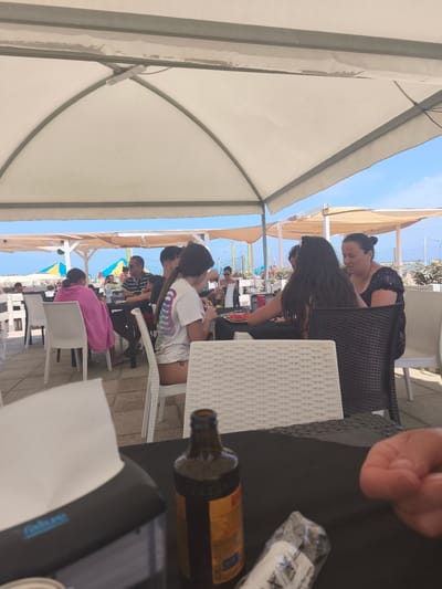The image shows a group of people sitting at tables under a large, white canopy at what appears to be an outdoor restaurant or beach cafe in San Salvo, Italy. In the foreground, a black table is partially visible with a tissue dispenser, a bottle of beer, and a wrapped item. A person's hand is also in the foreground, reaching towards the wrapped item.
Several individuals are seated at tables in the midground, some facing each other and appearing to converse. One person in the mid-left is wearing a pink towel draped over their shoulders, while another person in the mid-right is wearing a white t-shirt with a design on the back. The background reveals more tables and chairs, and beyond that, beach umbrellas and the sky. The overall setting suggests a casual, sunny day at a coastal establishment.