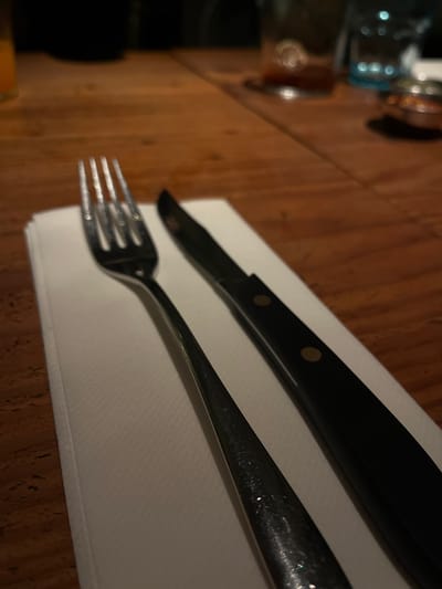 The image shows a place setting with a fork and knife on a white napkin atop a wooden table. The knife has a black handle with two visible rivets. The fork and knife are parallel and positioned diagonally across the napkin. A blurred bottle or glass is visible on the left side of the table. The location in the city of Barranco, Peru, cannot be determined from the image.