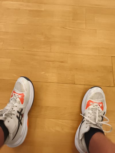 The image shows a pair of white and orange athletic shoes on a light brown wooden floor. The shoes have black accents, including the Asics logo on the side of each shoe. The word "METARISE" is visible in black text on the side of the left shoe. The shoes appear to be new and are tied with white laces. The scene is lit from above, suggesting an indoor setting with artificial lighting. The wooden floor has visible grain and appears to be polished. The perspective is from the wearer's point of view, looking down at their feet. There is no indication of the time of day or weather conditions. The location context is Pegnitz, Germany, but there are no specific visual cues in the image to confirm this. The image appears to be a still photograph.
