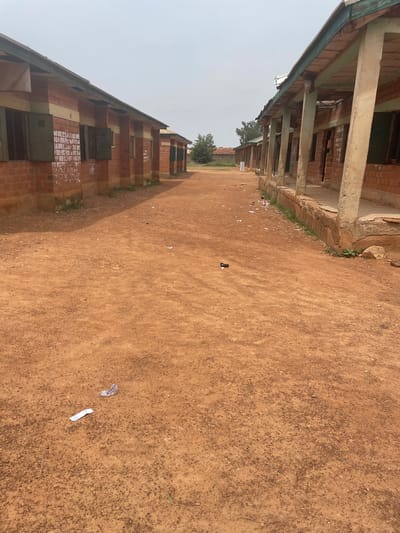 The image depicts an outdoor scene in Television, Nigeria, showing a pathway between two brick buildings. The pathway is covered in reddish-brown dirt, with scattered debris, including pieces of paper and a dark object that resembles a shoe or a piece of footwear.
The buildings appear to be school classrooms or similar structures. The building on the left is made of red brick with green-shuttered windows and shows some faded writing on its exterior walls. The building on the right has a covered walkway supported by concrete pillars, also constructed with red brick, and has visible signs of wear and tear. The sky above is overcast, suggesting a cloudy or hazy day. The overall environment appears to be a school compound or a similar institutional setting, with an unpaved ground surface and basic architectural structures.