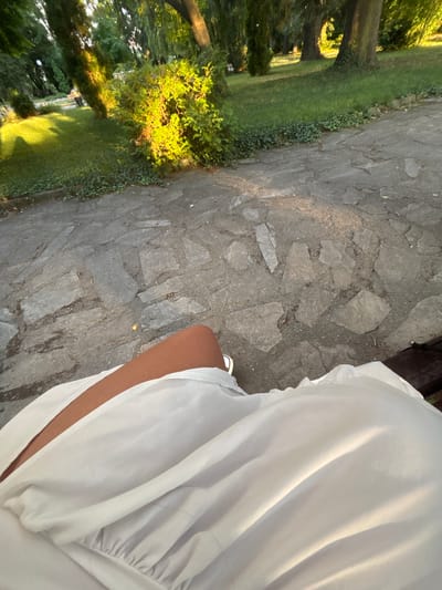 The image shows a person sitting on a bench in an outdoor setting, likely a park or garden. The foreground is dominated by the person's white clothing, which is draped over their lap and legs. A glimpse of the person's legs and a white sandal are visible. The background reveals a stone pathway, a grassy area with ivy, and several trees. The sunlight casts shadows and highlights on the scene. The location is identified as the town of Garmen, Bulgaria. There are no other individuals visible in the image, and the focus is on the person's attire and immediate surroundings. The scene is quiet and appears to be captured during daylight.