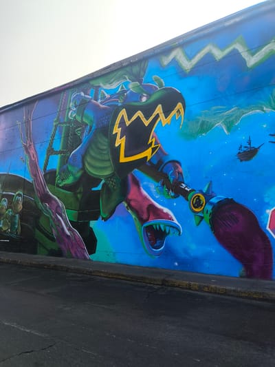 The image shows a mural on a wall, likely located in La Victoria, Peru. The mural features a cartoonish, monstrous figure with a reptilian or dragon-like head and body. It seems to be piloting a ship or a vehicle with mast-like structures visible behind it. The figure is holding a weapon or tool with a purple brush-like end. In the background, there is a bright blue sky with white, mountainous forms and a small, distant sailing ship. Two smaller cartoon figures are visible near the base of the masts of the ship, on the left. The mural's color palette is dominated by blues, greens, purples, and yellows. A paved road is visible in the foreground.