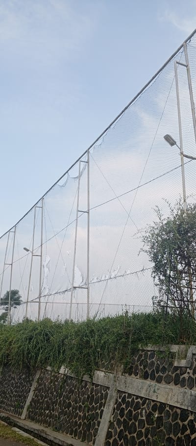 The image depicts a chain-link fence with visible damage, featuring several large tears and holes. The fence is supported by metal posts and a metal frame. A street lamp is attached to one of the vertical support posts. Green vegetation, appearing as dense bushes and vines, grows along the top of a retaining wall constructed from dark, irregular stones. The retaining wall has concrete supports. The sky is a pale blue with some wispy clouds. The scene appears to be outdoors, possibly in a park or sports facility, with a hint of urban infrastructure suggested by the street lamp and the retaining wall. There are no people or animals visible in the image. The overall condition of the fence suggests it has been exposed to wear and tear or damage. The location is specified as Kudawa, Sri Lanka, though this detail is not visually verifiable from the image content alone.