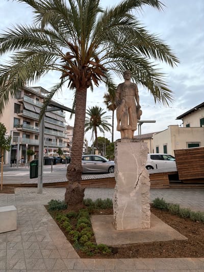The image depicts a public square in Port d'Alcúdia, Spain, with a statue of a man and a child in the foreground. The statue, made of stone, stands on a rough-hewn stone pedestal. The man is depicted wearing simple clothing and holding a hat. Behind the statue, a large palm tree dominates the scene, its fronds casting shadows. In the background, a modern building with balconies and awnings is visible, along with other palm trees and some parked cars. The ground is paved with textured tiles, and there are some wooden benches and planters with greenery. The sky is overcast. The overall impression is that of a pleasant, somewhat tropical urban environment.