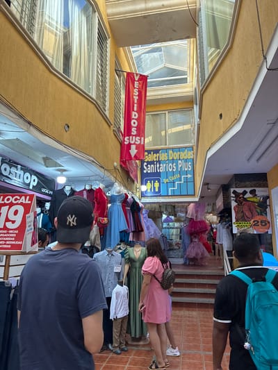 The image displays an indoor commercial pedestrian thoroughfare in Coatzacoalcos, Mexico, characterized by a narrow passage between multi-story buildings with textured yellow walls and a brownish-orange tiled floor. An overhead skylight structure provides natural illumination, supplemented by internal lighting fixtures.
Retail clothing stores line both sides of the passage. On the left, a shop with a black banner (partially obscured, possibly "oshoop") and a white-red sign indicating "LOCAL 19" displays items such as blouses, lycra, skirts, bermudas, and t-shirts. Mannequins in this shop exhibit a black shirt, a red shirt, and a blue dress. A vertical red banner reading "VESTIDOS SUSI" hangs prominently from the ceiling structure. On the right side, a blue sign indicates "Galerias Doradas Sanitarios Plus" with an arrow and restroom symbols, pointing towards an upper level accessible via a visible set of brown concrete steps. Shops beyond the stairs prominently display various formal dresses, including pink, purple, blue, and green garments, on mannequins and hangers. A large poster on the right wall advertises "PERIMEN CAMISAS JEANS" with a 25% discount, featuring a male model.
In the foreground, a male subject is observed from behind, wearing a dark blue t-shirt and a black baseball cap with a white "NY" logo. In the midground, a female subject walks away from the viewer, clad in a short pink dress and carrying a small patterned backpack. Adjacent to her, another person's lower legs are visible. To the right, a second male subject, also seen from behind, wears a black t-shirt with white trim and carries a bright teal backpack. Several other individuals are visible in the background, navigating the commercial space.