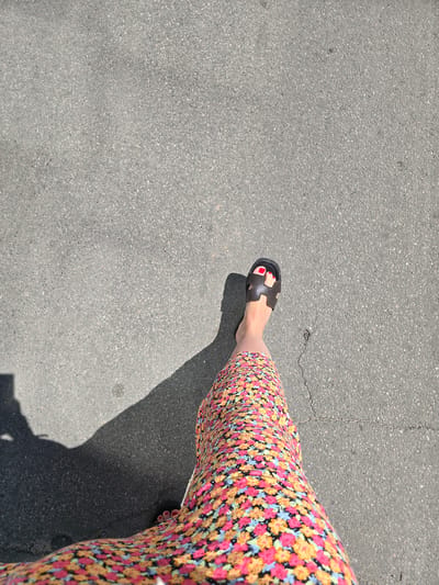 The media file is a top-down shot of a person's legs and feet walking on asphalt. The person is wearing a colorful, floral-print skirt and dark, flat sandals. Their toenails are painted red. The asphalt is gray and textured, with a few visible cracks and some dappled sunlight casting shadows. The scene appears to be outdoors on a sunny day. The location context provided is Petropavlivska Borshchahivka, Ukraine. There is no visible text in the image. The image captures a simple, everyday moment of walking.