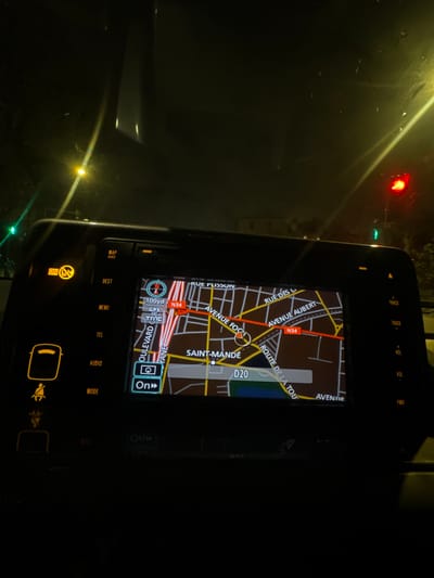 The image depicts a nighttime interior view from a vehicle, facing forward through the windshield. The central feature is a vehicle infotainment system displaying a navigation map. The map is centered on "SAINT-MANDÉ, France," clearly showing street names such as "RUE PLISSON," "AVENUE FOC," "AVENUE AUBERT," "BOULEVARD DE VANE," and "ROUTE DE LA TOU," along with routes "N34" and "D20." The GPS indicates a distance of "100yd" and shows active GPS and TMC services.
The infotainment console features illuminated physical buttons for "MAP VOICE," "DEST," "MENU," "TEL," "AUDIO," and "MODE" on the left, and "TRACK," "VOL," and "PWR" on the right. Two yellow warning indicators are lit: one representing a passenger seatbelt or airbag status, and another circular symbol, possibly for traction control.
Through the windshield, a green traffic light is visible to the left, and a red traffic light is visible to the right. Several yellow streetlights illuminate the dark exterior, casting reflections and light streaks on the windshield, indicating wetness or fog. Faint silhouettes of distant buildings are discernible against the dark sky. No individuals are visible in the scene.