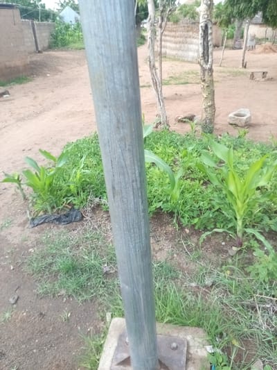 A medium shot captures a metallic pole in the foreground, positioned vertically and slightly to the left of the frame. The pole is weathered and shows visible vertical grain. Behind the pole, the scene opens to a dusty, unpaved area with sparse vegetation. To the left, a brick wall made of rough, tan-colored blocks forms a boundary. Beyond this wall, several structures are visible, including one with a light-colored, conical roof.
Further into the background, a cluster of young corn plants are growing in a patch of earth, interspersed with other green foliage. A black plastic bag lies on the ground near these plants. To the right of the corn, a dead tree trunk with a hollowed-out section stands prominently. The ground is predominantly dry, reddish-brown earth. In the far background, traditional dwellings with thatched roofs are visible, along with a few scattered trees. The overall environment suggests a rural or semi-urban setting, consistent with locations like Zinna, Nigeria. No people are present in the image.