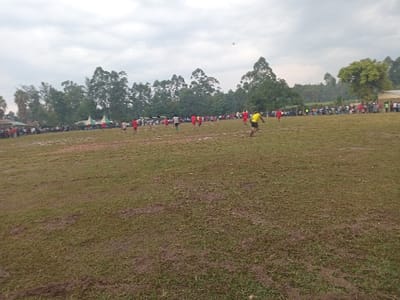 A soccer game unfolds on a muddy field under a cloudy sky. Several players in red and one in yellow are scattered across the field. A crowd lines the perimeter, observing the match. Behind the spectators, trees form a backdrop, and a few tents are visible to the left.