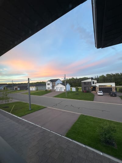 The image depicts a suburban street scene in Dieppe, Canada, during what appears to be twilight. The sky is a gradient of pale blue, pink, and orange, indicating sunset or sunrise. Several modern residential houses line the street, characterized by dark roofs, light-colored siding, and attached garages. Two cars are parked in the driveway of one prominent house, which features a mix of dark gray and white exterior elements, as well as stone accents near the entrance. The street itself is paved and shows some visible cracks. Lawns are well-maintained and green, with a few small trees planted in some yards. Overhead, power lines and a utility pole are visible. The foreground of the image is dominated by the angled perspective of dark-colored roof shingles, suggesting the photo was taken from an elevated viewpoint, possibly from a window or balcony.